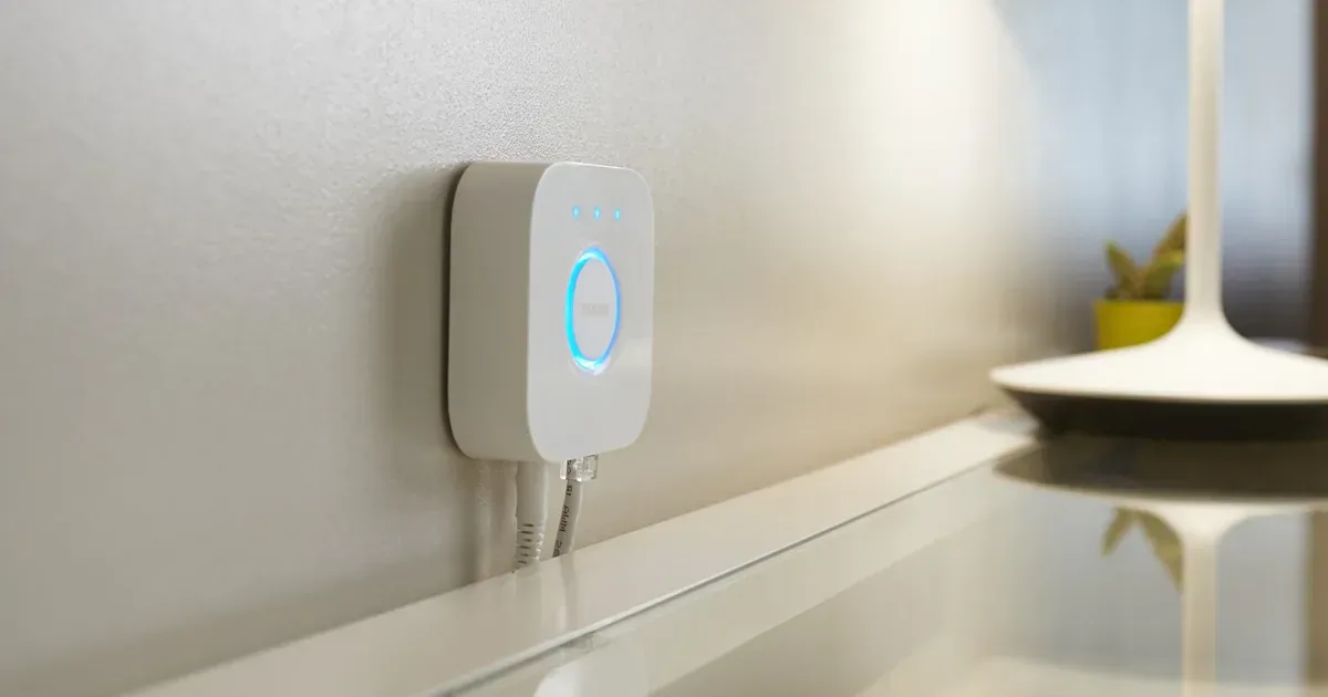 A white square Philips Hue Bridge mounted on a wall above a glass shelf, with a table lamp in the background