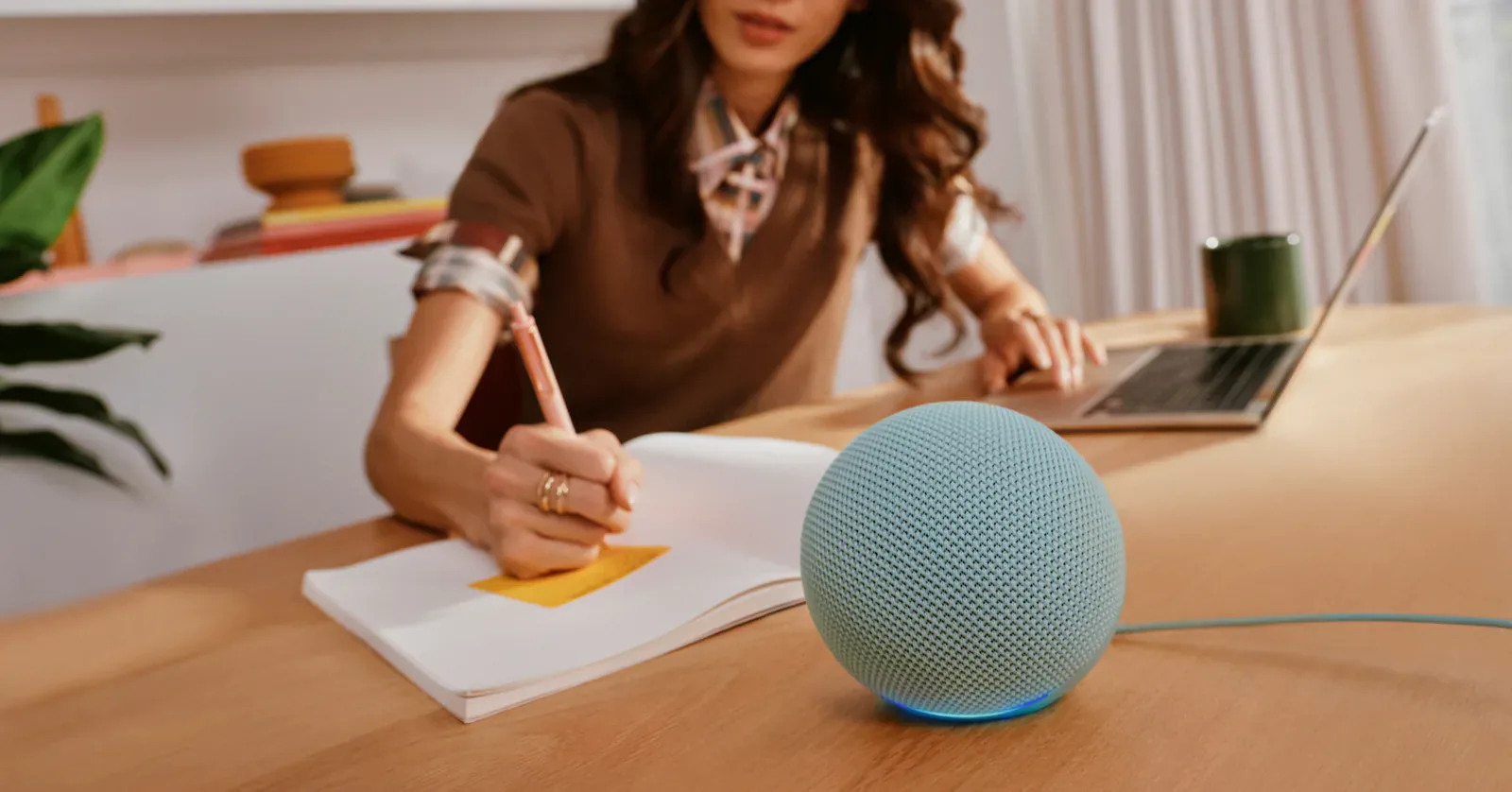 New Amazon Echo Dot in a blue-green fabric finish on a wooden desk while a person writes in a notebook behind it